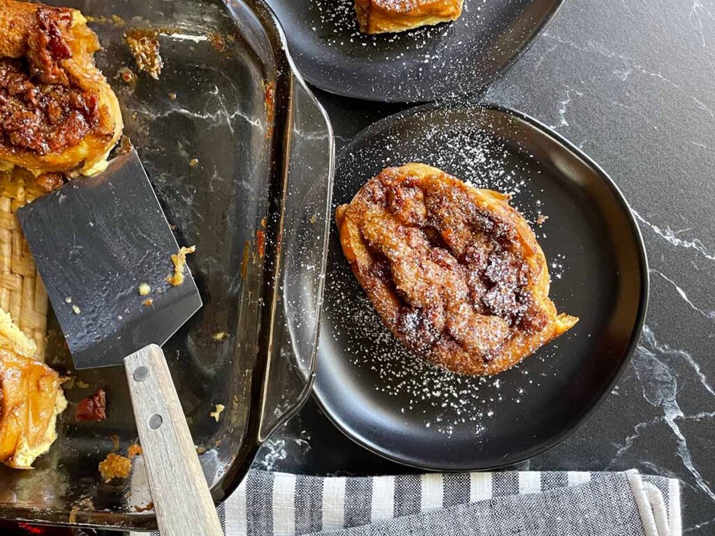 Slices of overnight maple bacon baked french toast on black plates garnished with powdered sugar.